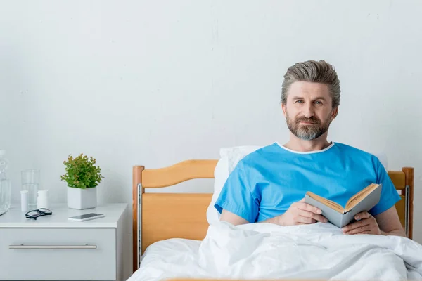 Paciente en vestido médico sosteniendo libro y mirando a la cámara en el hospital - foto de stock