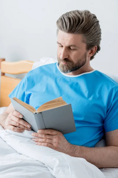 Guapo paciente en vestido médico libro de lectura en el hospital - foto de stock