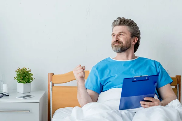 Patient heureux en robe médicale tenant presse-papiers et montrant un geste oui à l'hôpital — Photo de stock