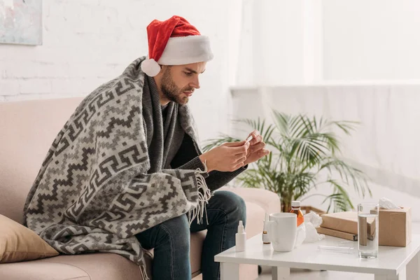 Sick man in santa hat, wrapped in blanket, sitting near table with medicines and holding thermometer — Stock Photo