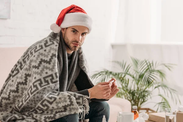 Kranker Mann mit Weihnachtsmütze, in Decke gehüllt, blickt in die Kamera, während er das Thermometer hält — Stockfoto