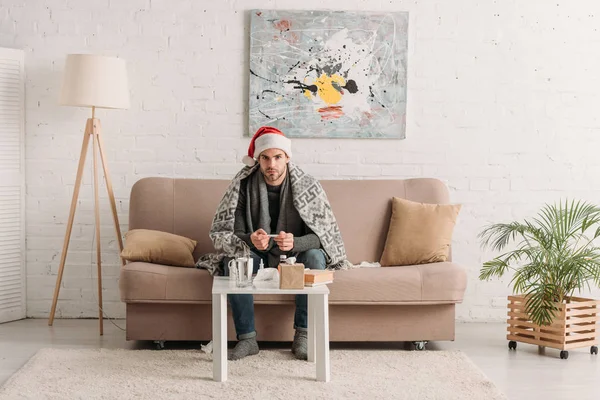 Kranker Mann mit Weihnachtsmütze, in Decke gehüllt, sitzt auf Sofa neben Tisch mit Medikamenten — Stockfoto