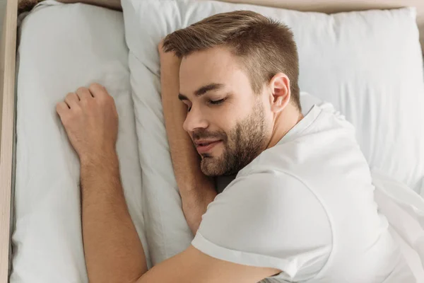 Ansicht von oben: junger Mann lächelt, während er auf weißem Bettzeug schläft — Stockfoto