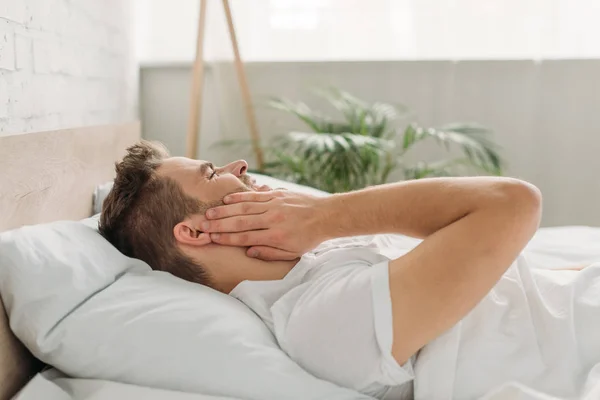 Joven tocando la mejilla mientras está acostado en la cama y sufriendo de dolor de muelas - foto de stock