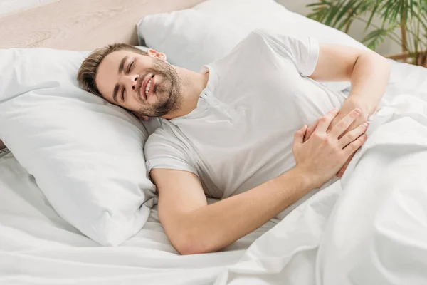 Joven acostado en la cama y sufriendo de dolor de estómago con los ojos cerrados - foto de stock