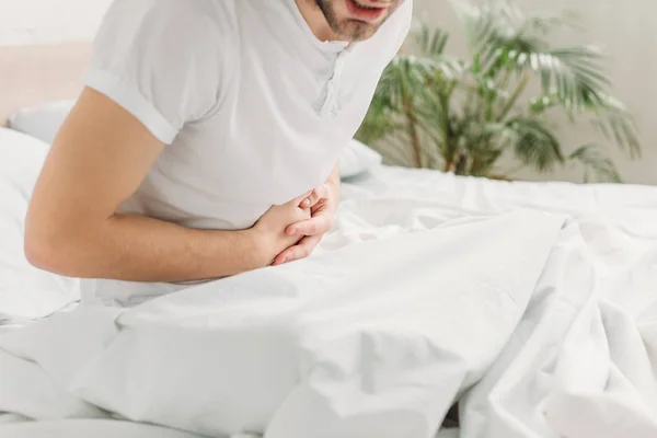 Vista recortada del hombre sentado en la cama y que sufre de dolor abdominal - foto de stock