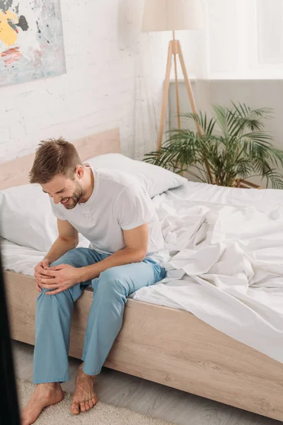 Молодой человек, сидящий на кровати и страдающий от боли в колене — стоковое фото