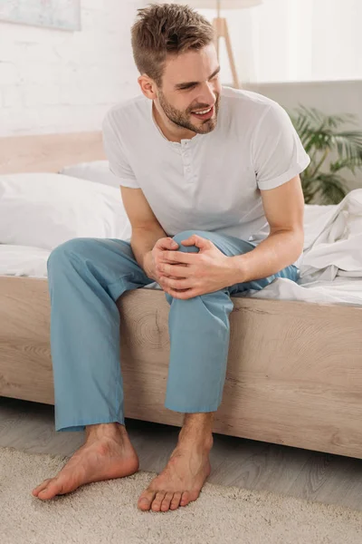 Joven tocando la rodilla mientras está sentado en la cama y sufriendo de dolor - foto de stock