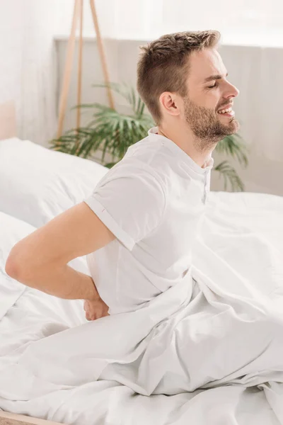 Joven sentado sobre ropa de cama blanca y sufriendo de dolor en el lomo con los ojos cerrados - foto de stock