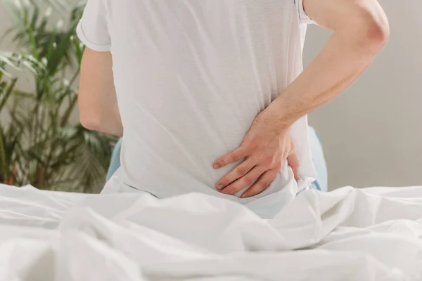 Vista ritagliata di uomo che tocca lombata mentre seduto sul letto e soffre di dolore — Foto stock