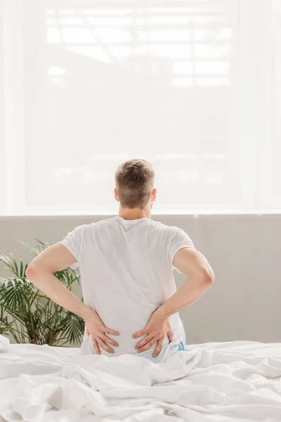 Вид сзади человека, который касается спины, сидя на кровати и страдая от боли — стоковое фото