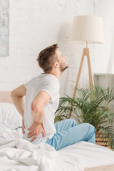 Junger Mann sitzt auf weißem Bettzeug und leidet unter Rückenschmerzen — Stockfoto