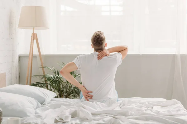 Rückenansicht eines Mannes, der auf weißem Bettzeug sitzt und unter Rückenschmerzen leidet — Stockfoto