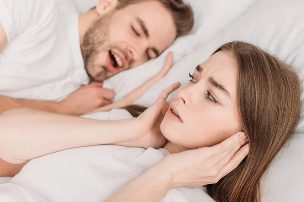 Selektiver Fokus der Frau, die Ohren mit den Händen verstopft, während sie in der Nähe des schnarchenden Ehemanns im Bett liegt — Stockfoto