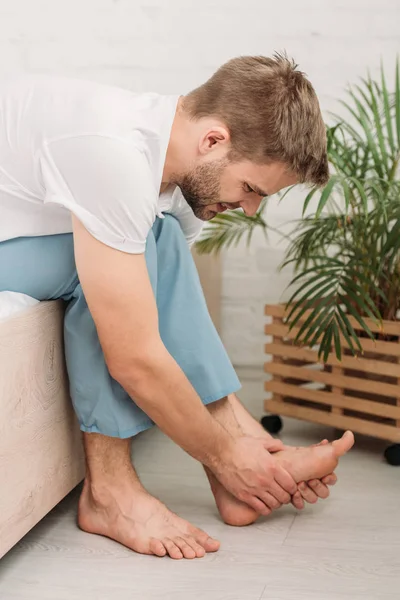 Jeune homme touchant le pied tout en étant assis sur le lit et souffrant de douleur — Photo de stock