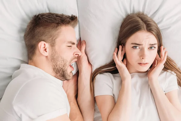 Vue de dessus de la femme insatisfaite bouchant les oreilles avec les mains et regardant la caméra tout en étant couché dans le lit près de ronflement mari — Photo de stock