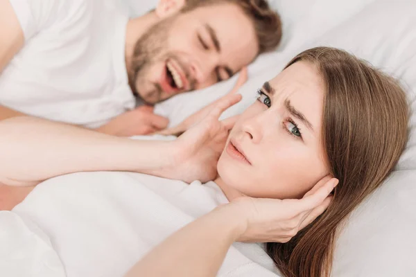 Dissatisfied woman plugging ears with hands and looking at camera while lying in bed near snoring husband — Stock Photo