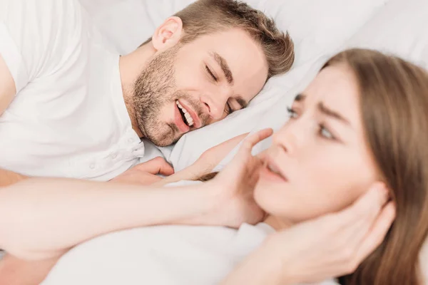 Selective focus of exhausted woman plugging ears with hands while lying in bed near snoring husband — Stock Photo
