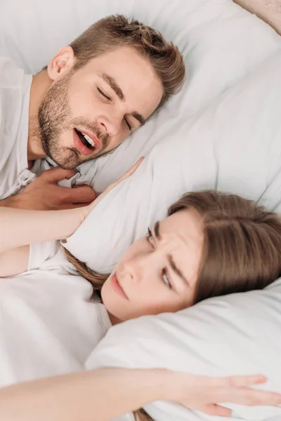 Awakened woman plugging ears with pillow while lying in bed near snoring husband — Stock Photo