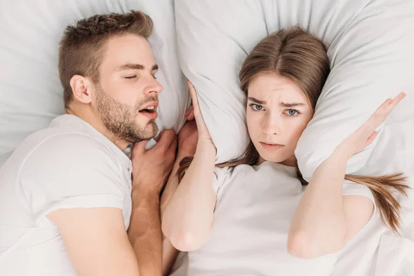 Top view of woman plugging ears with pillow while lying in bed near snoring husband — Stock Photo