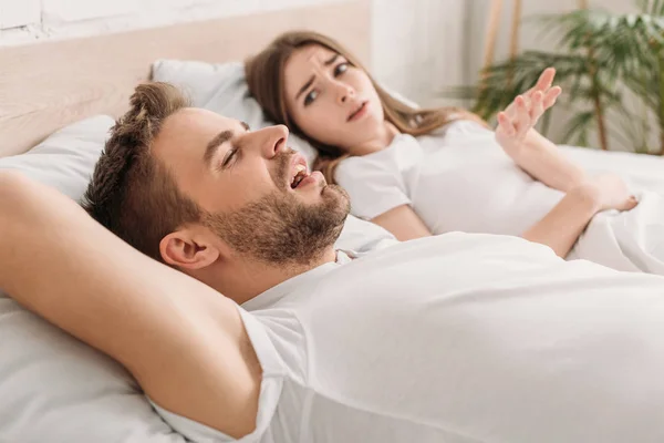 Незадоволена жінка демонструє обурливий жест, лежачи в ліжку біля хропіння чоловіка — стокове фото
