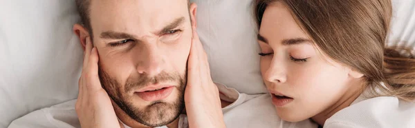 Top view of exhausted man plugging ears with hands while lying in bed near snoring wife, panoramic shot — Stock Photo