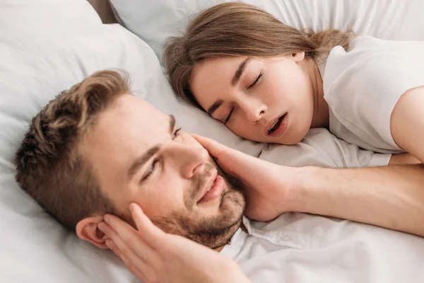 Виснажений чоловік, що вимикає вуха руками, лежачи в ліжку біля хропіння дружини — стокове фото