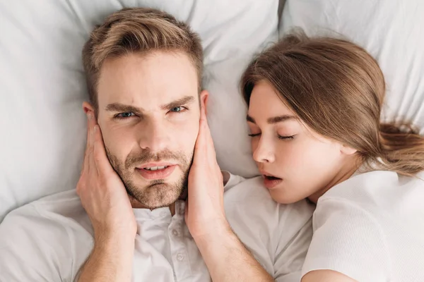 Homem descontente tapando ouvidos com as mãos e olhando para a câmera enquanto deitado na cama perto de ronco esposa — Fotografia de Stock