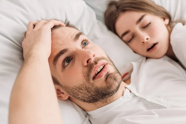 Проснувшийся мужчина, держась за голову, лежа в постели рядом с храпящей женой — стоковое фото