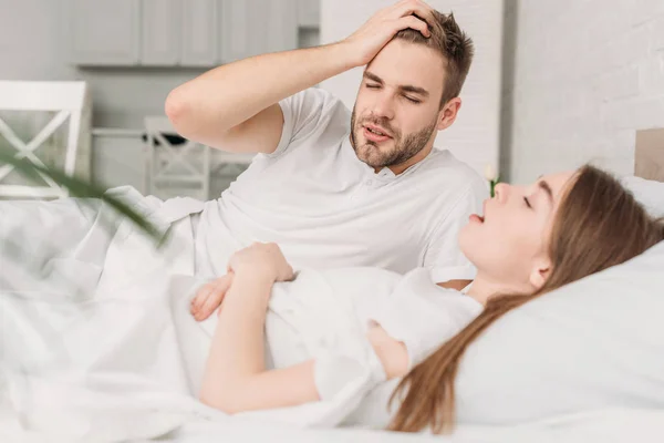 Erschöpfter Mann hält Hand auf Kopf, während er neben schnarchender Frau im Bett liegt — Stockfoto