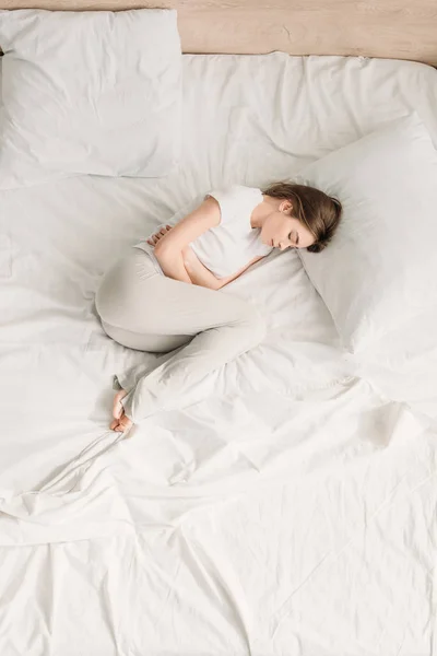 Vista superior de la niña acostada en la cama con los ojos cerrados y que sufre de dolor abdominal - foto de stock