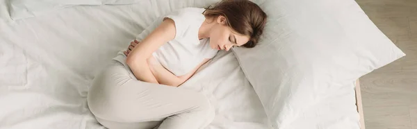 Top view of young woman lying in bed and suffering from stomach pain, panoramic shot — Stock Photo