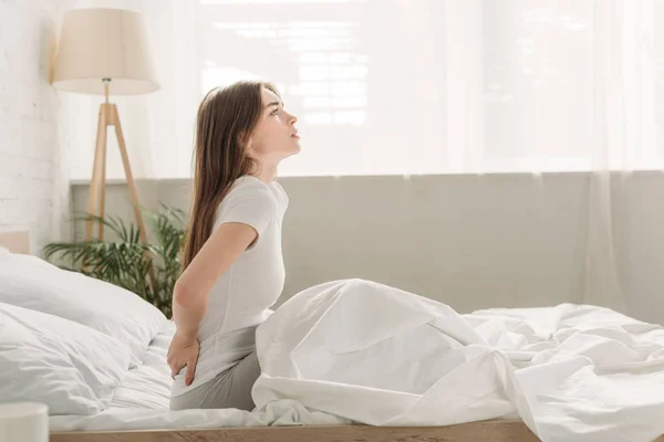 Side view of upset girl looking away while sitting in bed and suffering from back pain — Stock Photo