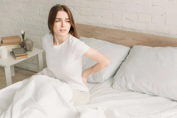 Aufgebrachtes Mädchen schaut weg, während es im Bett sitzt und unter Lendenschmerzen leidet — Stockfoto
