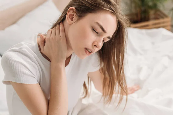 Jovem mulher sentada na cama com os olhos fechados e sofrendo de dor no pescoço — Fotografia de Stock