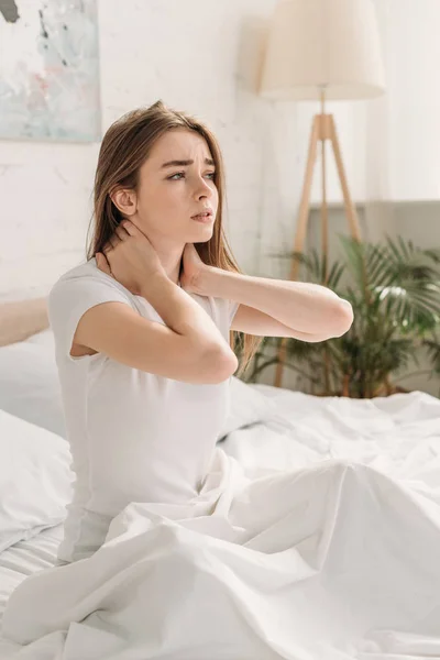 Upset girl looking away while sitting in bed and suffering from neck pain — Stock Photo