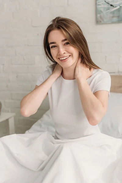 Cheerful young woman smiling at camera while sitting in bed and holding hands on neck — Stock Photo