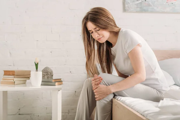 Frowned girl sitting on bed near bedside table and suffering from knee pain — Stock Photo