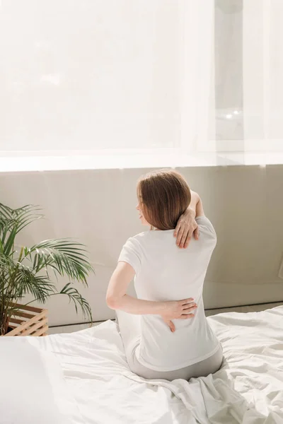 Vista posterior de la chica sentada en la cama y que sufre de dolor de espalda - foto de stock