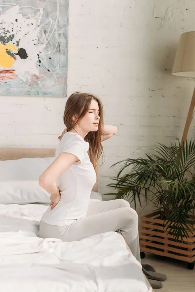 Menina bonita sentada na cama com os olhos fechados, enquanto sofre de dor nas costas — Fotografia de Stock