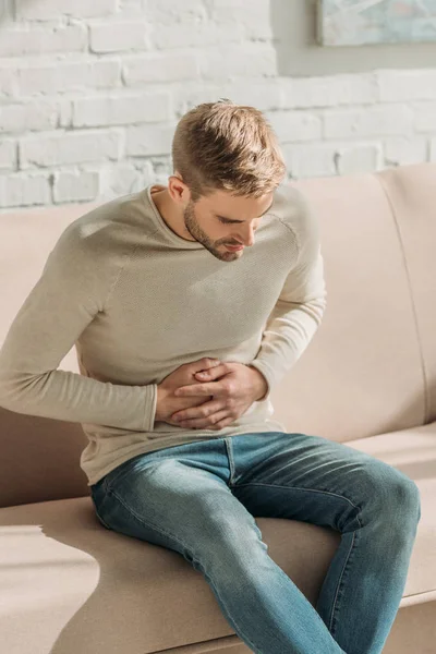 Young man sitting on sofa at home and suffering from stomach pain — Stock Photo