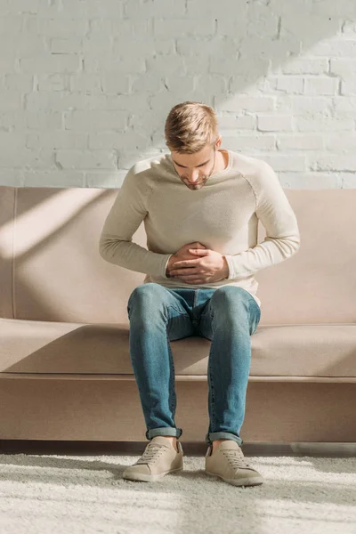 Joven sentado en el sofá y tocando el estómago mientras sufre de dolor abdominal - foto de stock