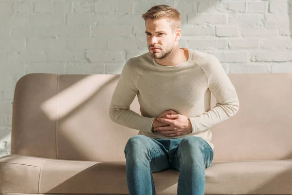 Upset man sitting on sofa and touching stomach while suffering from abdominal pain — Stock Photo