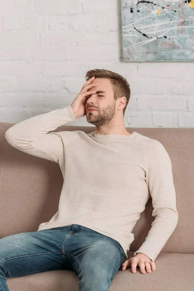 Young man sitting on sofa with closed eyes while suffering from migraine — Stock Photo