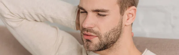 Panoramic shot of man with closed eyes sitting on sofa and suffering from neck pain — Stock Photo