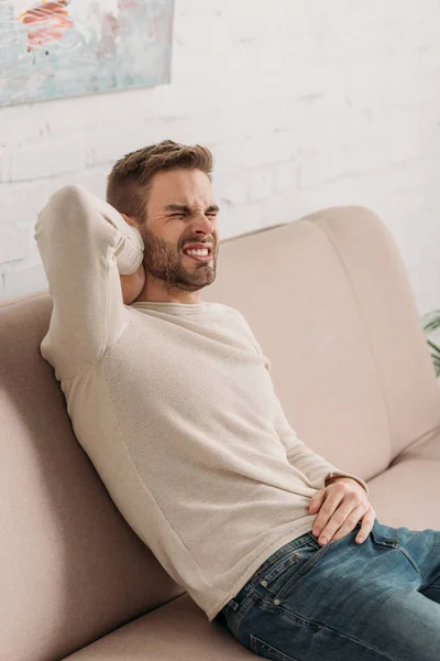 Jeune homme assis sur le canapé et souffrant de douleur au cou — Photo de stock