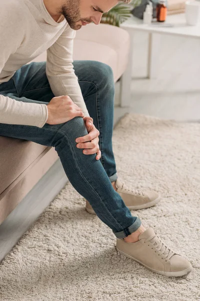 Young man sitting on sofa and touching knee while suffering from pain — Stock Photo