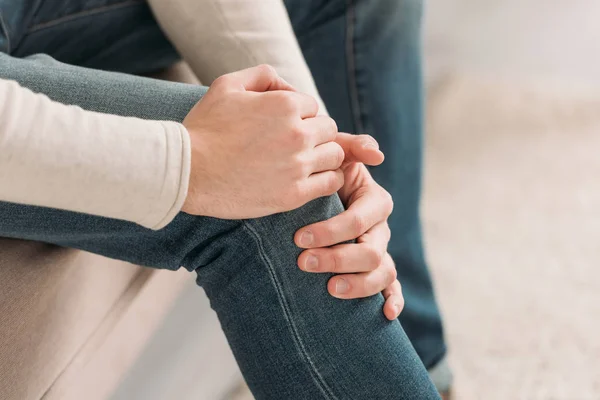 Cropped view of man sitting on sofa and touching knee while suffering from pain — Stock Photo