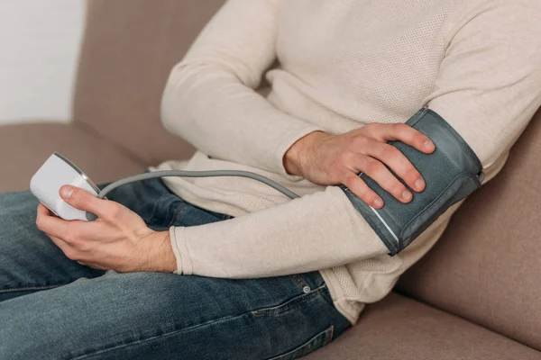 Обрезанный вид человека, сидящего на диване и измеряющего артериальное давление тонометром — стоковое фото