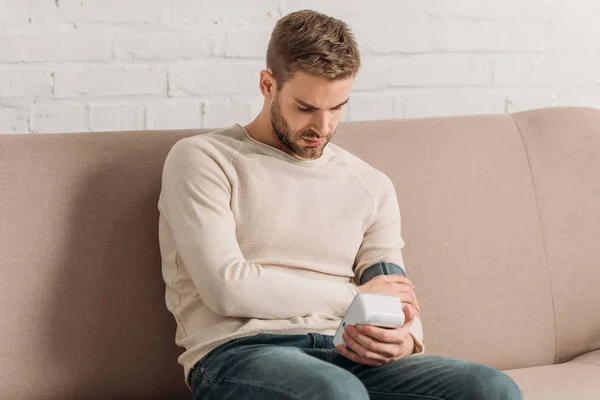 Young man sitting on sofa and measuring blood pressure with tonometer — Stock Photo